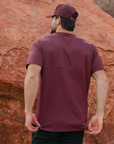 Man wearing a maroon t-shirt and cap standing against a rocky background